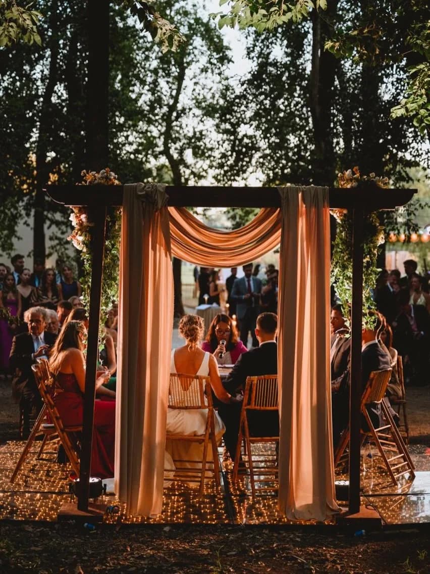 Ceremonia al atardecer bajo una pérgola de madera con telas claras, flores y guirnaldas de luces entre los árboles.